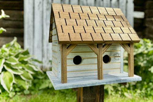 A handcrafted wooden Mini Martin birdhouse with a shingled roof and dual entry holes, mounted outdoors on a post with a soft garden background.