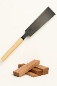 Japanese Ryoba pull saw with dual-edge blade and rattan-wrapped handle, displayed above stacked hardwood blocks on a clean light background.